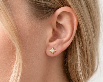 May include: A gold stud earring shaped like a multi-pointed star with a small, clear, round gemstone set in the centre. The star features sharp, radiating points and a polished metallic finish, secured firmly against the earlobe.
