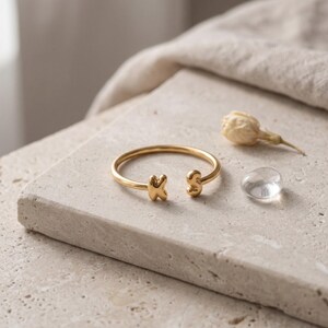 May include: A gold-toned open ring featuring two rounded, puffed-style letters, K and S, at each end of the band. The ring has a smooth, polished finish and a circular shape, designed to be worn on a finger with the lettered ends facing outward.