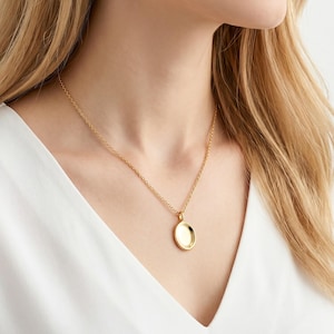 Puede incluir: Un collar dorado con una cadena fina y un pequeño colgante ovalado. El colgante tiene un diseño simple y pulido que refleja la luz. El collar se lleva sobre una blusa blanca. La joya es un accesorio clásico.