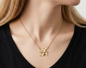 Könnte beinhalten: Eine goldfarbene Halskette mit einem Anhänger in Form eines vierflügeligen Windrads. Der Anhänger hängt an einer zarten, passenden goldfarbenen Kette. Das Design ist poliert und reflektierend und fängt das Licht durch seine flachen Flächen ein.