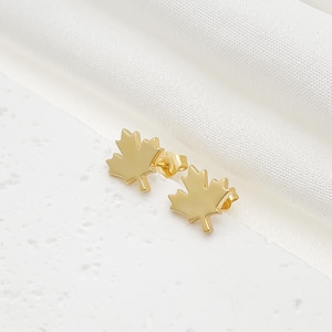 Könnte beinhalten: Ein Paar goldfarbene Ohrstecker in Form von Ahornblättern. Die Ohrringe werden auf einer weißen, strukturierten Oberfläche präsentiert. Die Ohrringe sind klein und zart.