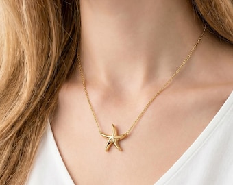 May include: A gold-toned necklace featuring a polished starfish pendant suspended from a delicate, thin chain. The starfish has five smooth, curved arms and a reflective metallic finish, resting against a plain white fabric neckline.