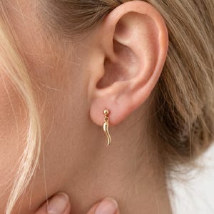 May include: A close-up of a gold-colored earring. The earring features a small ball at the top and a dangling horn-shaped charm. The earring is worn on a person's ear.