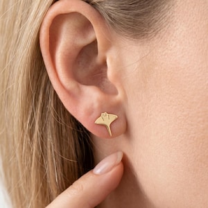 Puede incluir: Pendientes de botón dorados con forma de manta raya. Los pendientes presentan un diseño detallado del cuerpo y la cola de la raya. Los pendientes se muestran en una oreja, destacando su tamaño y estilo.