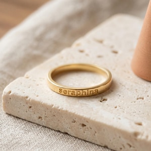 Puede incluir: Un anillo de metal dorado con un acabado liso y pulido. La superficie exterior de la banda presenta el nombre seraphina grabado en letras minúsculas sin remates. El anillo tiene un diseño circular sencillo con un ancho constante.