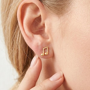 May include: A close-up of a gold-colored stud earring in the shape of a musical note. The earring is worn on a person's ear. The musical note has a rounded bottom and a square top. The earring is shiny and reflects light.