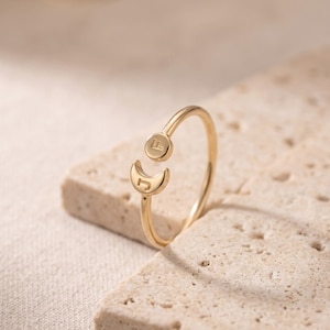 Puede incluir: Un anillo abierto de tono dorado que presenta un pequeño dije circular grabado con la letra F y un dije de media luna grabado con la letra J. La banda es delgada y pulida, creando un diseño delicado y sencillo para el dedo.
