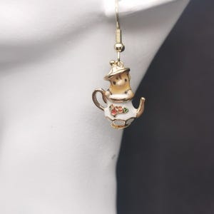 Puede incluir: Un pendiente dorado con forma de tetera, con cuerpo blanco, tapa y asa marrones. La tetera tiene detalles florales rojos y verdes y está suspendida de un alambre dorado. Un accesorio caprichoso.