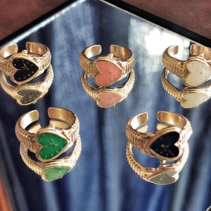 May include: Six gold-toned adjustable rings with heart-shaped faces in black, pink, white, green, and navy blue. The rings are displayed on a reflective surface, showcasing their intricate design and color variations.