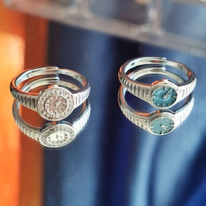 May include: Two silver-toned rings with watch face designs. One ring features a light-colored face with clear stones around the perimeter. The other ring has a teal-colored watch face. Both rings are displayed on a reflective surface.