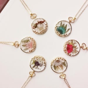 Puede incluir: Una colección de delicados collares con colgantes redondos. Cada colgante presenta una disposición diferente de flores secas encapsuladas en resina transparente, engastadas en un bisel dorado. Los collares tienen cadenas finas de color plateado.
