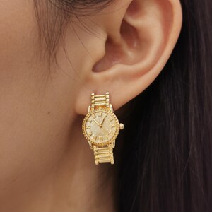May include: Gold-colored watch earrings. The earrings are shaped like miniature watches, with a round face featuring Roman numerals and a textured band. The watch earrings are attached to the ear with a post.