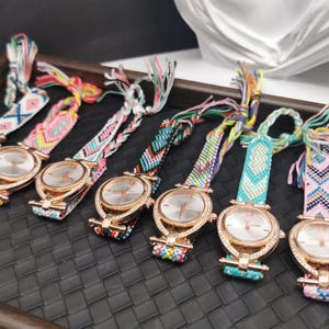 May include: A collection of seven rose gold watches with woven, colorful bands. The watch faces are silver with delicate hands. The bands feature various geometric patterns in shades of pink, blue, green, and yellow.