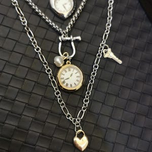 May include: A multi-layered necklace with silver chains and various pendants. The pendants include a heart-shaped watch, a round watch with gold detailing, a key, and a heart-shaped lock. The backdrop is a textured black surface.