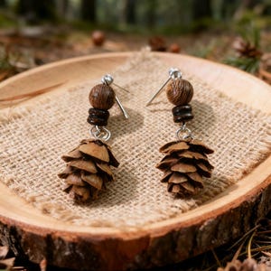 Puede incluir: Pendientes hechos a mano con colgantes de piñas naturales. Cada pendiente tiene un alambre para la oreja plateado, cuentas de madera y una pequeña piña. Los pendientes se exhiben sobre una rodaja de madera con un fondo de arpillera.