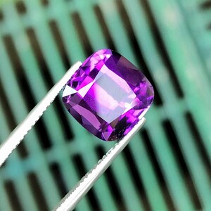 Può includere: Una gemma viola scuro, tagliata quadrata e sfaccettata, è tenuta da una pinzetta d'argento. La gemma ha una lucentezza brillante, che riflette la luce. Lo sfondo è sfocato, verde e nero.