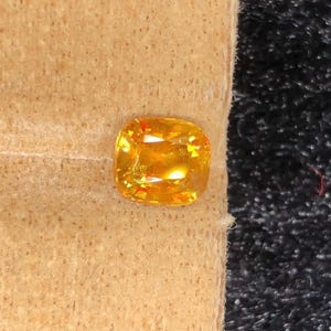 Può includere: Una gemma giallo dorato, taglio a cuscino, poggia su una superficie beige strutturata. La gemma ha una superficie luminosa e riflettente, su uno sfondo scuro e strutturato. È probabile che si tratti di uno zaffiro o di un topazio.