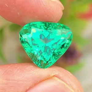 May include: A faceted, heart-shaped emerald gemstone. The gem is a vibrant green color and is held between two fingers. The facets create a sparkling effect, reflecting light. The background is blurred, with hints of green and purple.