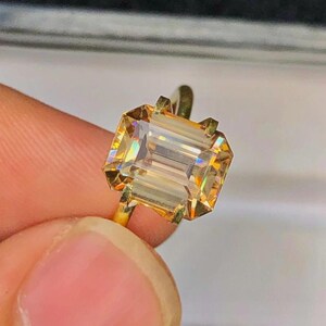 May include: A gold ring featuring a large, rectangular, light-yellow gemstone. The gemstone has a faceted cut, reflecting light with a rainbow effect. The ring's band is a simple, polished gold.
