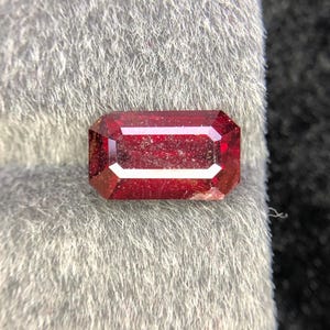 Può includere: Una gemma rettangolare sfaccettata di colore rosso intenso. La pietra è posta su uno sfondo morbido e grigio, dalla consistenza vellutata. Il taglio della pietra rivela molteplici sfaccettature, esaltandone la brillantezza e la chiarezza. Potrebbe essere un rubino o una pietra preziosa simile.