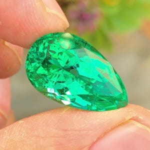 Può includere: Una gemma di smeraldo sfaccettata a forma di pera. La gemma è di un colore verde vibrante ed è tenuta tra due dita. Il taglio della pietra crea molteplici superfici riflettenti, esaltandone la brillantezza. Lo sfondo è sfocato, con sfumature di verde e viola.