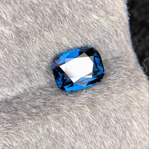 May include: A rectangular, faceted gemstone with a deep blue hue. The gem is set against a soft, grey, textured background. The facets reflect light, creating a brilliant sparkle. This gemstone is likely for jewellery.