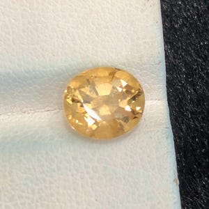 May include: Oval-cut, golden-yellow gemstone. The gem is set against a white leather-like surface, with a dark, textured background. The gemstone's facets reflect light, creating a sparkling effect. This gemstone is suitable for jewelry making.