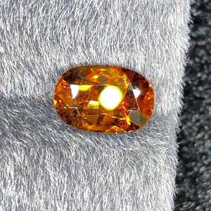 Può includere: Una gemma ovale arancione dorata con un punto luce centrale brillante. La gemma è posta su uno sfondo morbido e grigio testurizzato, creando un contrasto che ne esalta la brillantezza. Le sfaccettature della gemma riflettono la luce, mostrando la sua chiarezza e il colore.
