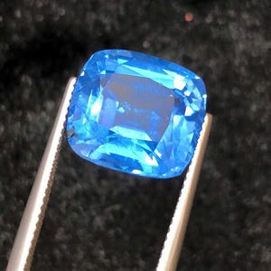Könnte beinhalten: Ein leuchtend blauer, kissenförmig geschliffener Edelstein, der von einer Pinzette vor einem dunklen Hintergrund gehalten wird. Die Facetten des Edelsteins reflektieren das Licht und zeigen seine Klarheit und Farbe. Die Pinzette ist silberfarben.