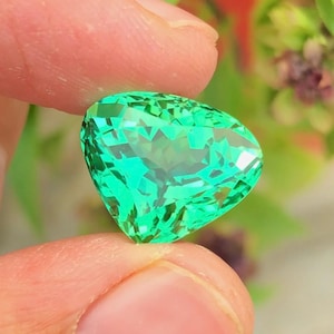 Può includere: Una gemma sfaccettata a forma di cuore, in una tonalità verde brillante e vibrante. La gemma è tenuta tra due dita, mostrando i suoi intricati tagli e le superfici riflettenti. Lo sfondo presenta un fogliame verde sfocato.