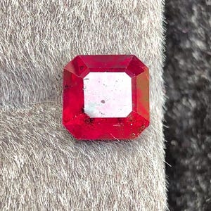 Può includere: Una gemma rosso scuro sfaccettata, tagliata a forma rettangolare, con un centro più chiaro di forma quadrata. La gemma è posta su uno sfondo grigio strutturato, con uno sfondo grigio scuro.