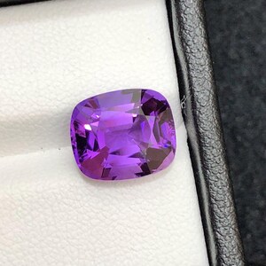 Può includere: Una gemma di ametista sfaccettata a taglio cuscino. La gemma è di colore viola intenso ed è posta su uno sfondo bianco all'interno di una scatola nera. Le sfaccettature creano un gioco di luci e ombre.