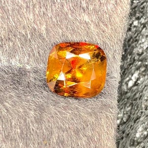 Può includere: Una gemma sfaccettata, taglio a cuscino, con una calda tonalità arancione dorato. La gemma mostra riflessi interni ed è posta su uno sfondo grigio tenue. Il taglio della gemma mette in risalto la sua brillantezza e chiarezza.