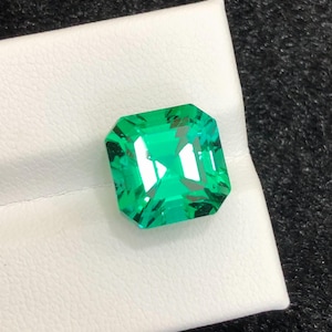 Può includere: Una pietra preziosa di colore verde brillante con taglio quadrato e superficie sfaccettata che riflette la luce. La pietra presenta bordi netti e puliti e un aspetto trasparente e brillante, con una profonda tonalità smeraldo.