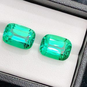 May include: Two loose, rectangular, cushion-cut gemstones with a vibrant, translucent green colour. These faceted stones feature sharp, clear edges and a polished surface, displayed side-by-side within a white-lined, black jewellery box.