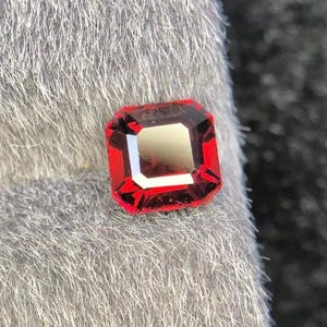 Può includere: Una gemma sfaccettata, taglio rettangolare, di un rosso vibrante con un centro più chiaro. La gemma è posta su uno sfondo grigio, strutturato, che ne esalta la brillantezza. Adatta per gioielli.