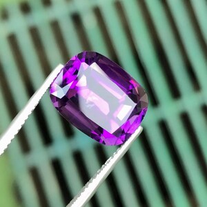Può includere: Una gemma di ametista rettangolare e sfaccettata tenuta da una pinzetta. La gemma è di un colore viola intenso con riflessi interni. Lo sfondo è un motivo sfocato verde e nero.