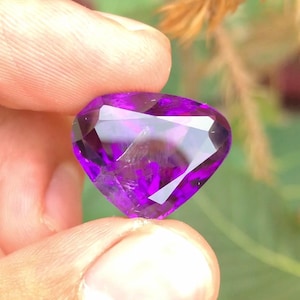 May include: A faceted, heart-shaped amethyst gemstone. The gem is a deep purple color with internal inclusions and a brilliant cut. The stone is held between two fingers against a blurred green and brown background.