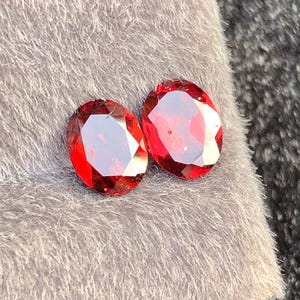 Può includere: Due gemme rosse, tagliate a forma ovale e sfaccettate. Le gemme sono esposte su una superficie grigia e strutturata. Le gemme hanno una qualità brillante e riflettente, che mette in risalto le loro sfaccettature interne e il colore vivace.