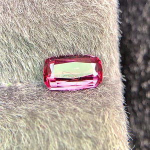 Può includere: Una gemma rettangolare sfaccettata di colore rosa acceso. La pietra ha bordi leggermente arrotondati ed è posta su uno sfondo grigio strutturato. Le sfaccettature catturano la luce, creando un effetto scintillante. La gemma sembra essere una pietra tagliata.