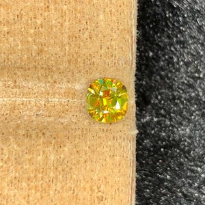 May include: A vibrant, cushion-cut gemstone with a brilliant yellow hue. The gem is set against a light brown, textured surface, with a dark gray background. The facets of the stone reflect light, creating a sparkling effect.