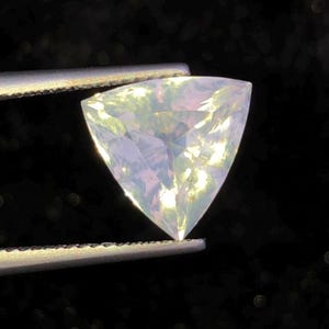 May include: A triangular, iridescent gemstone held by tweezers against a dark background. The gem has a pale, opalescent color with hints of yellow and purple. The facets catch the light, creating a shimmering effect. This could be a loose gemstone for jewelry making.