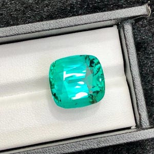 May include: A cushion-cut gemstone with a vibrant, translucent teal-green colour and faceted surfaces that reflect light. The gemstone is displayed inside a black rectangular box with a white interior lining, showcasing its clarity and colour.