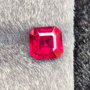 May include: A vibrant, faceted, square-cut ruby gemstone. The gem is a deep, saturated red color, with sharp edges and a polished surface. The ruby is set against a soft, gray, textured background.