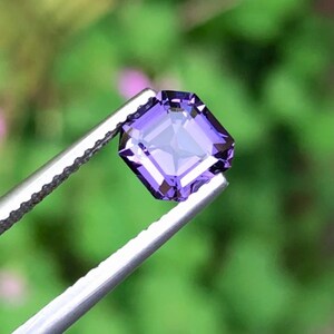 以下が含まれることがあります： 銀色のピンセットで保持された、正方形にカットされた紫色の宝石。 宝石のファセットは光を反射する対称的なパターンを作り出しています。 ピンセットは、ぼやけた緑色の背景に対して鮮明に焦点を当てています。 宝石は鮮やかな紫の色合いです。