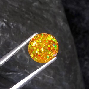 May include: A round, faceted gemstone held by silver tweezers. The gem is a vibrant yellow with flashes of green and red, showcasing a brilliant cut. The background is a dark, textured surface.