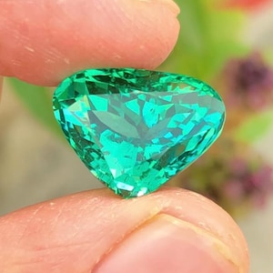 Può includere: Una gemma sfaccettata a forma di cuore, di un vibrante colore verde smeraldo. La gemma è tenuta tra due dita, mostrando il suo taglio intricato e la sua brillantezza. Lo sfondo è sfocato, con accenni di fogliame verde e viola.