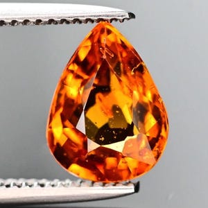 Peut inclure: Une pierre précieuse en forme de goutte d'eau, facettée, tenue par une pince argentée. La gemme est d'une couleur orange vif, avec des facettes internes réfléchissant la lumière. Le fond est gris doux.