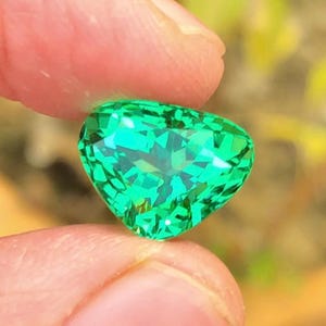 May include: A heart-shaped, faceted, bright green gemstone is held between two fingers. The gem has a brilliant cut, reflecting light and creating a sparkling effect. The background is blurred, with hints of green and yellow.