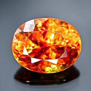 May include: A faceted, oval-cut gemstone displaying a vibrant mix of orange, yellow, and red tones. The gem's internal structure reflects light, creating a brilliant display. The gemstone is set against a dark background, highlighting its vibrant colours.